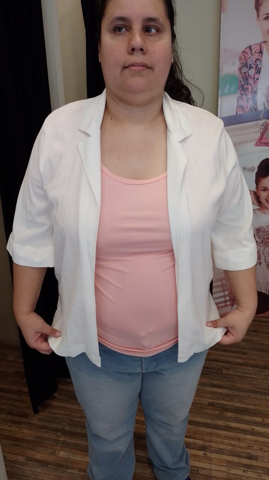 Una mujer de tez morena clara está de pie y mira directamente al frente con una expresión serena, casi estoica. Su rostro es de forma redonda, con cejas oscuras y espesas que enmarcan sus ojos marrones. Su nariz es ancha y sus labios son de tamaño medio, con un tono rosado natural. Tiene el cabello oscuro, recogido hacia atrás, dejando ver parte de su frente.