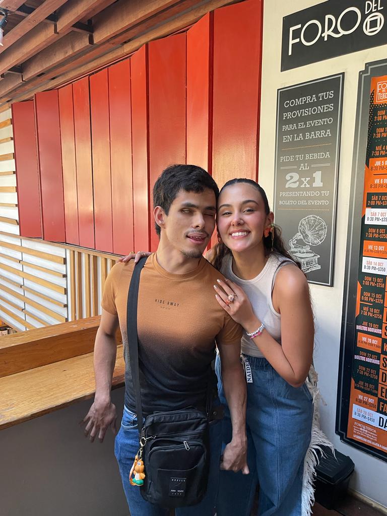 Soy Ale, parado aquí junto a Susana. Mi pelo castaño oscuro es corto, y siento mi barba incipiente en mi piel de tono medio. Entrecierro los ojos, sacando un poco la lengua, un gesto tonto, tal vez. Llevo mi camiseta degradada, que se oscurece desde un marrón óxido a negro, con "HIDE AWAY" escrito. La bandolera negra está sobre mi hombro, con ese llavero infantil colgando.  Susana es radiante. Su pelo castaño oscuro está recogido, dejando su cara clara y sonriente a la vista.